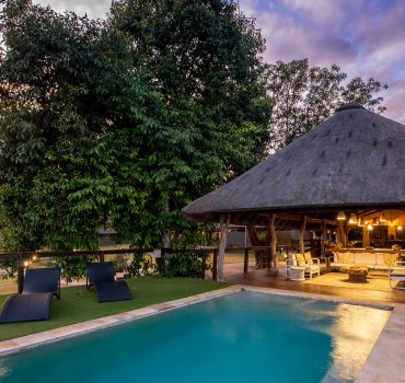 Outdoor pool and lounge area at Arathusa Safari Lodge with lush greenery and a thatched-roof structure.