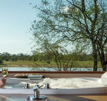 Luxurious bathtub with a view at Arathusa Safari Lodge, featuring a woman relaxing with a glass of champagne, overlooking the serene wilderness and a tranquil waterbody.